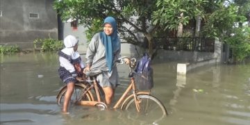 Seorang ibu hendak mengantarkan anaknya pergi sekolah di tengah banjir. Foto: Bacaini/Syailendra