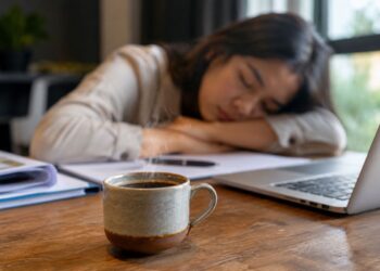 Seseorang memegang secangkir kopi sambil mengantuk saat bekerja di depan laptop