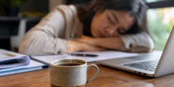 Seseorang memegang secangkir kopi sambil mengantuk saat bekerja di depan laptop