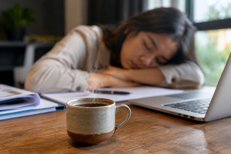 Seseorang memegang secangkir kopi sambil mengantuk saat bekerja di depan laptop